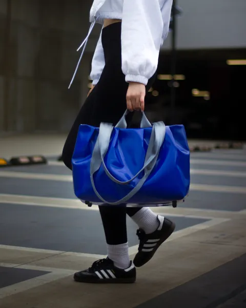 A person is carrying a blue, shiny backpack over their shoulder and is in motion. They are dressed in a white coat, black tights, white socks, and black Adidas shoes. The background features a crosswalk and a slightly blurred urban environment.