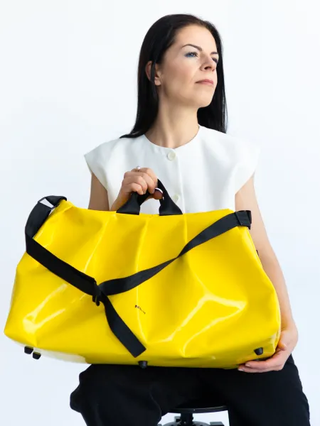 A person is holding a large, yellow backpack with black straps and a black belt around the middle. They are wearing a white top and black pants. The background is light and plain, drawing attention to the backpack and the person.