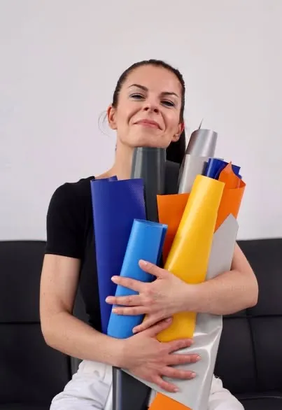 A smiling person is holding several large, colorful rolls in their arms, which come in various colors like blue, orange, gray, and white. They are wearing a black top and are sitting on a black couch. The background is light and plain.