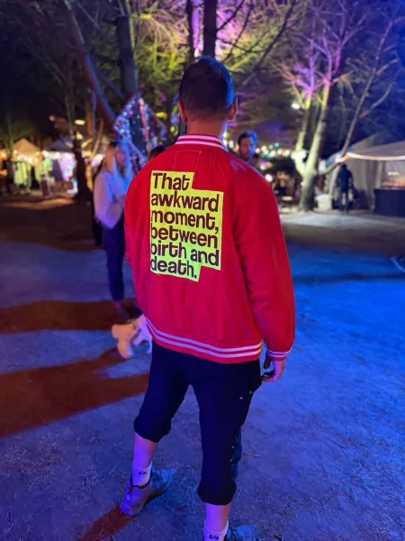 Eine Person steht von hinten fotografiert in einem beleuchteten Park. Sie trägt eine rote Jacke mit einem gelben Aufdruck auf dem Rücken, der lautet: "That awkward moment, between birth and death." Im Hintergrund sind weitere Menschen und beleuchtete Bäume zu sehen.