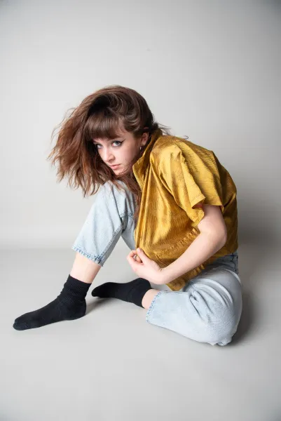 Eine Person mit langen, braunen Haaren und einem blassen Teint sitzt auf dem Boden, während sie eine goldene Bluse und eine hellblaue Jecke trägt. Sie hat schwarze Socken an und hält ihren Arm in einer entspannten Pose.