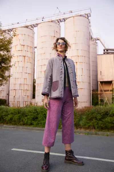 A person stands in front of an industrial facility with large, rusty silos. They are wearing a patterned jacket in gray and beige with large pockets, matching wide pants in purple, and sturdy boots. The person is wearing sunglasses and appears confident.