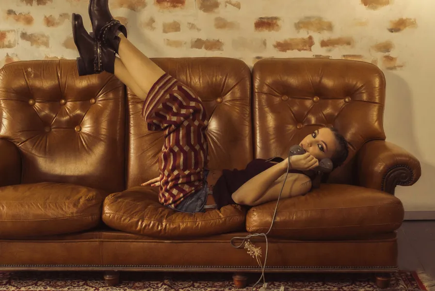 A person is lying relaxed on a brown leather sofa with padded armrests and patterned wallpaper in the background. They are wearing a colorful, patterned shirt, jeans, and black boots while holding a microphone and looking into the camera. The atmosphere feels casual and stylish.