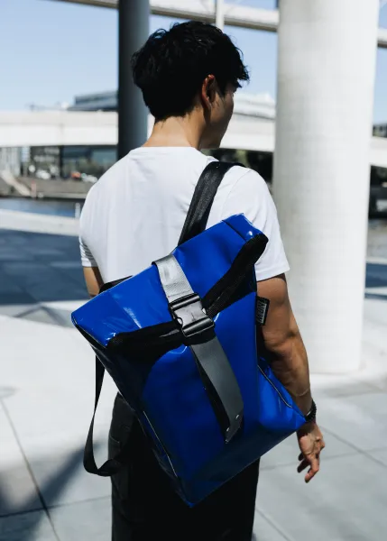 A person is carrying a blue, shiny backpack over their shoulders and walking through an urban environment. They are wearing a white t-shirt and black pants. The background features modern architecture with glass and concrete, as well as a slightly blurred street.