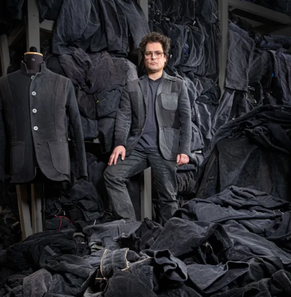 Ein Mann steht in einem Raum, der mit verschiedenen Kleidungsstücken und Stoffen in dunklen Tönen gefüllt ist. Er trägt eine graue Jacke mit großen Taschen und eine Brille. Im Hintergrund sind weitere Kleidungsstücke und ein Mannequin zu sehen, das ebenfalls mit dunkler Kleidung bedeckt ist.