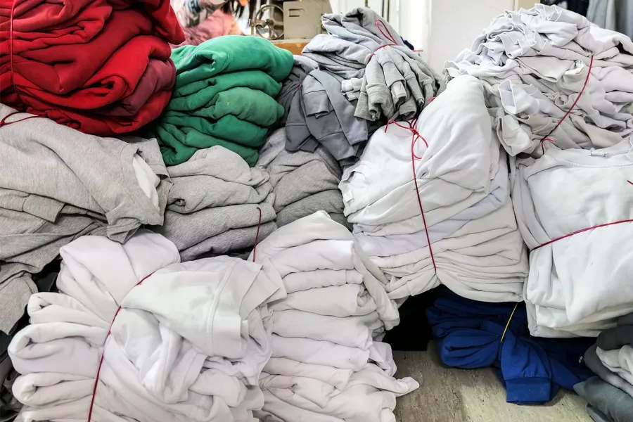 Several stacks of clothing in various colors, including red, green, gray, and white, are neatly bundled and tied together with red strings. The clothing items are arranged in a room with a gray floor and a white background. Additional clothing items and a gray cabinet can be seen in the background.