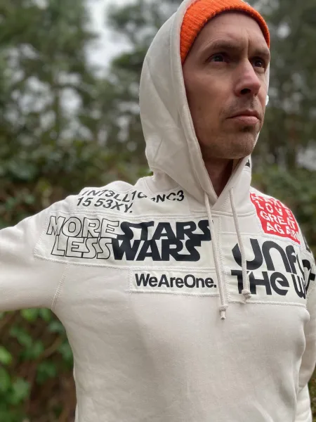 Ein Mann trägt eine helle Kapuzenjacke mit verschiedenen Aufdrucken. Auf der Schulter steht prominent "MORE STARS LESS WARS" in großen, fett gedruckten Buchstaben. Im Hintergrund sind grüne Bäume und ein leicht bewölkter Himmel zu sehen.