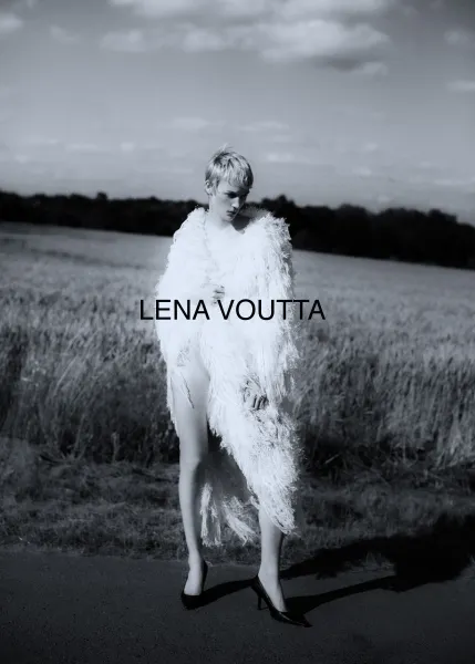 A person with short, light blonde hair stands in a wide, grassy field wearing a striking, white, fluffy outfit that resembles feathers. The sky is cloudy, and the person is wearing black high heels. The name "LENA VOUTTA" is prominently displayed in the center of the image.