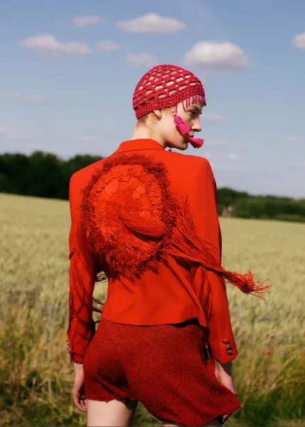A person stands in a wide, grassy field wearing a striking, red outfit consisting of a jacket with large, decorated feathers on the back and a short skirt. The person also wears a red, patterned beanie and a pink accessory on the ear. The sky is clear with some clouds, and the person looks into the distance.