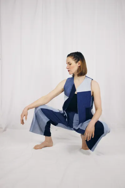 A woman strikes a fluid, dance-like pose against a minimalist backdrop of white fabric. Her lower body is crouched, with one leg slightly lifted, while her arms and torso lean gracefully forward, creating a sense of dynamic movement. She wears a sleeveless, asymmetric jumpsuit composed of bold geometric patches in black and light blue, combining rigid angular shapes with fluid draping. 