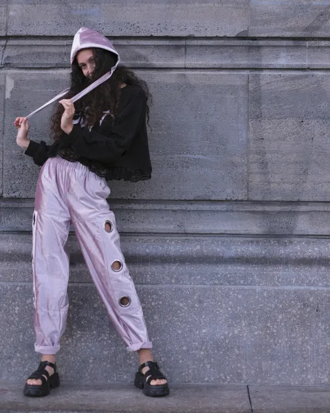 A person is posing on a wide, textured stone staircase with a gray stone wall in the background. They are wearing a long, dark-colored hoodie with a distressed lace overlay at the hem, paired with oversized, pink satin pants adorned with multiple silver-cap eyelets along the outer seams. The pants are loose-fitting and slightly rolled at the hems.