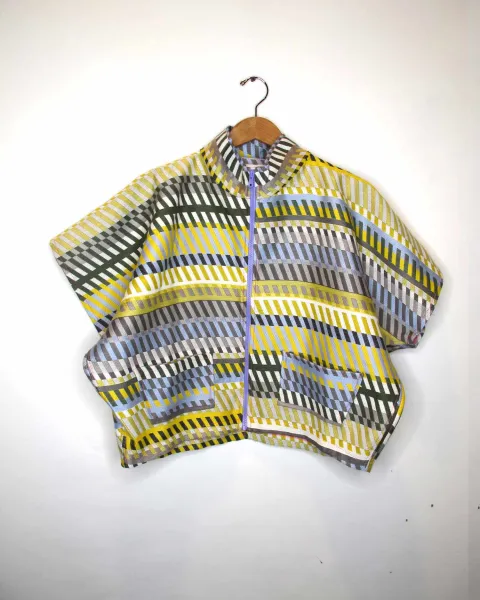 Ein Poncho mit einem auffälligen Muster aus gelben, grauen, weißen und blauen Streifen und Quadraten, der an einem Holzbügel hängt. Der Poncho hat einen hohen Kragen und ist vor einem hellen Hintergrund zu sehen.