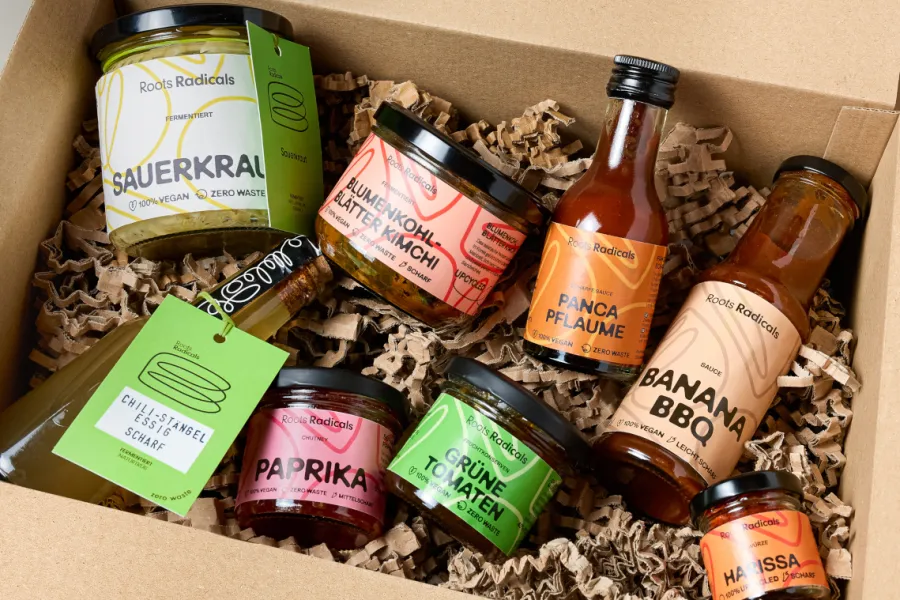 A box is filled with various fermented foods and spices packaged in jars and bottles. The products have labels with names like "Sauerkraut", "Cauliflower Leaf Kimchi", "Panca Plum", "Banana BBQ", "Green Tomatoes", "Paprika", "Chili Stem Vinegar", and "Harissa". The packaging comes in various colors and emphasizes the vegan and zero-waste philosophy of the brand Roots Radicals.