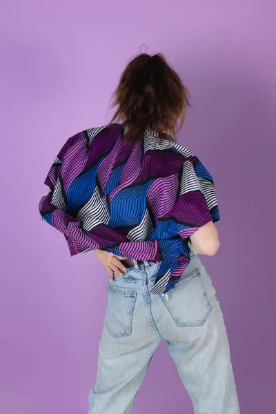 Eine Person steht mit dem Rücken zur Kamera und trägt einen Poncho mit einem lebendigen Muster aus pinken, blauen, schwarzen und weißen Wellenformen. Der Poncho ist weit geschnitten und wird mit hellblauen Jeans kombiniert. Der Hintergrund ist lila.