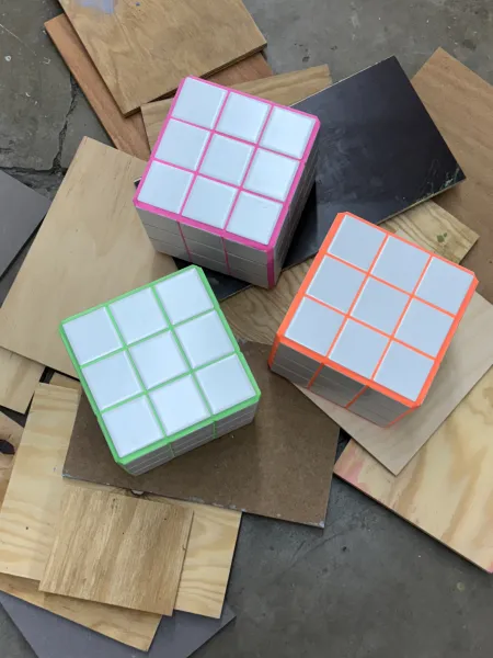Drei bunte Würfel mit den Farben pink, grün und orange liegen auf einem Holzboden, umgeben von verschiedenen Holzplatten. Die Zauberwürfel sind teilweise gelöst und zeigen ihre weißen Innenquadrate.