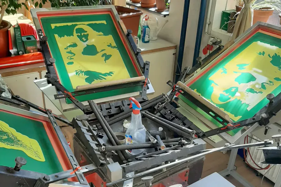 Eine Siebdruckmaschine mit mehreren Schablonen, die ein grünes und gelbes Design aufweisen, ist zu sehen. Die Schablonen sind auf Rahmen befestigt und zeigen ein Motiv mit einem Gesicht und Blättern. Verschiedene Werkzeuge und Flaschen mit Flüssigkeiten sind um die Maschine herum platziert.