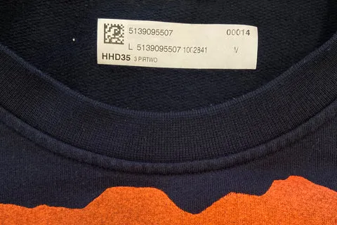 Ein dunkles Stoffstück mit einem orangefarbenen Saum ist zu sehen, das mit einem Etikett versehen ist. Das Etikett enthält einen QR-Code, eine Produktnummer, eine Größe und andere Identifikationsdetails.