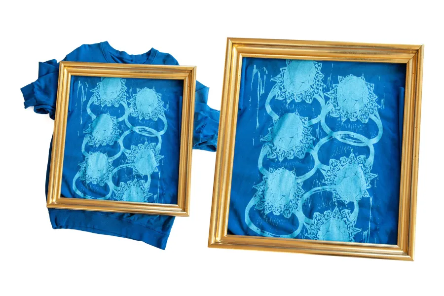 Zwei gerahmte Bilder zeigen ein blaues T-Shirt mit einem auffälligen Druck von ineinander verschlungenen Ringen und Sternen. Die Ringe sind mit detaillierten Mustern und Schriftzügen verziert, die an Graffiti erinnern. Die goldenen Rahmen heben die blauen Motive auf dem T-Shirt kontrastreich hervor.