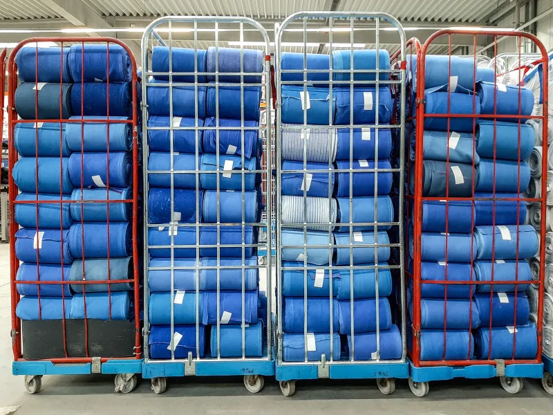 Three wire carts are loaded with rolled-up blue carpets neatly stacked in multiple rows. The carpets are wrapped in plastic and labeled, indicating a storage or recycling environment.