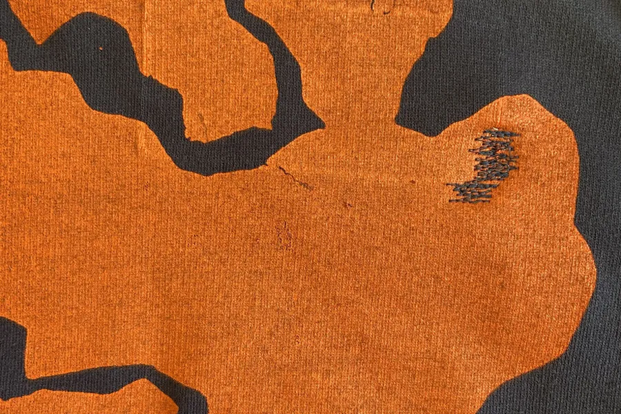 A piece of dark fabric with an orange hem is visible, tagged with a label. The label includes a QR code, a product number, a size, and other identification details.