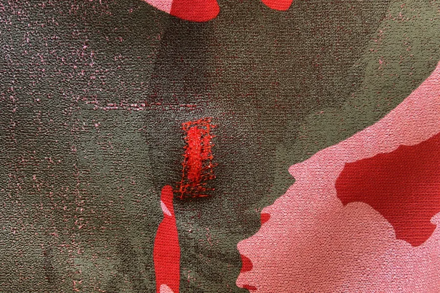 A piece of fabric displays a pattern of large, red flowers on a dark green background. One of the flowers is adorned with a small, embroidered text that reads "Spuren."