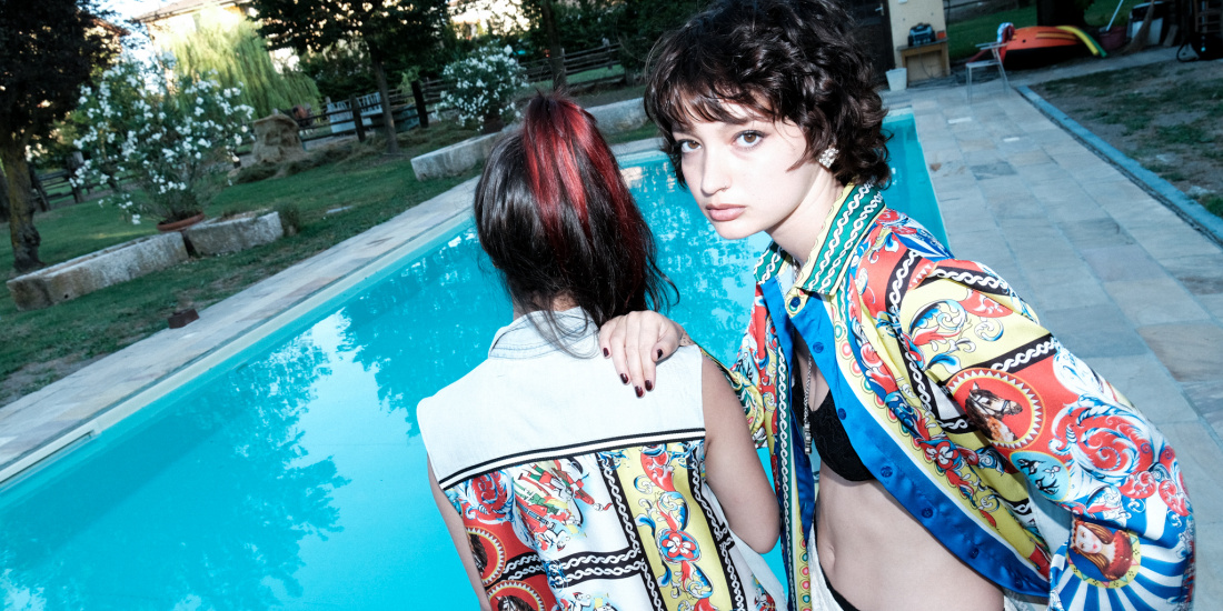 Two people are standing at the edge of a turquoise pool in a garden with trees and flowers. The person in the foreground is wearing a colorful, patterned shirt and a scarf, while the other person with red-streaked hair is wearing a white top. The atmosphere feels relaxed and summery.