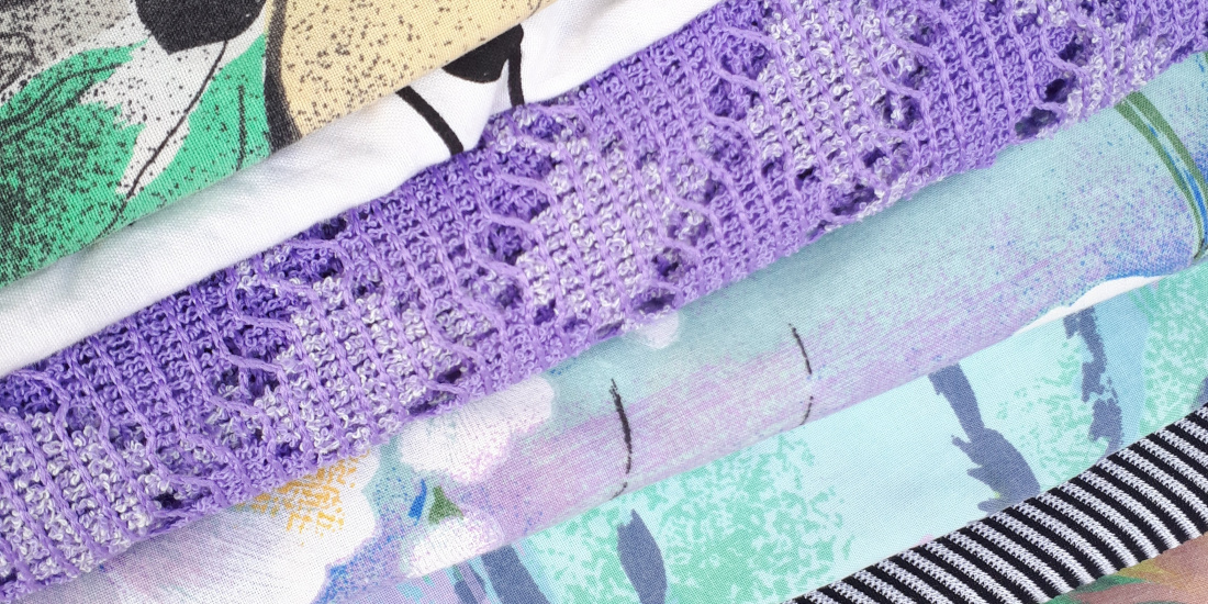 Various fabrics in vibrant colors and patterns are stacked on top of each other. The fabrics feature a mix of geometric and floral designs, including a purple fabric with a floral pattern, a black and white striped fabric, and a fabric with abstract patterns in green and red. The textures and colors of the fabrics are inviting and creative.