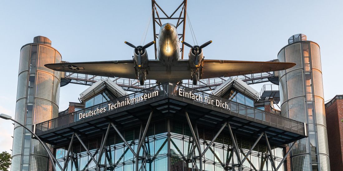 Ein großes Flugzeug ist auf dem Dach des Deutschen Technikmuseums in Berlin ausgestellt. Das Museum hat eine moderne Architektur mit großen Glasfenstern und zwei markanten, zylindrischen Türmen. Auf dem Dach steht der Schriftzug "Deutsches Technikmuseum Einfach für Dich."