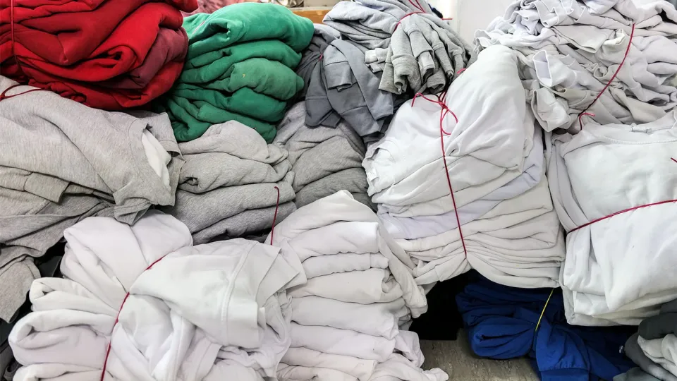 Several stacks of clothing in various colors, including red, green, gray, and white, are neatly bundled and tied together with red strings. The clothing items are arranged in a room with a gray floor and a white background. Additional clothing items and a gray cabinet can be seen in the background.