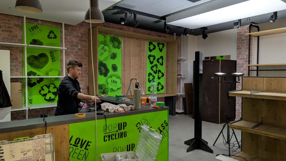 A man stands behind a green counter in a room with a cozy atmosphere. The counter is decorated with various items such as a water bottle, a thermos, and some cups. The walls are adorned with green posters featuring texts like "LOVE GREEN", "POPUP CYCLING", and "SMILE GREEN". The room has a mix of wooden and brick walls and is furnished with various pieces of furniture and lamps.