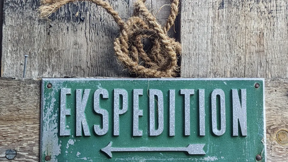 Ein Holzschild mit der Aufschrift "EKSPEDITION" in großen, grauen Buchstaben ist an einer Holzwand befestigt. Darüber hängt ein dekorativer Knoten aus Hanfseil. Das Schild hat eine grüne Farbe mit einigen abblätternden Stellen und ist mit vier Schrauben an der Wand befestigt.